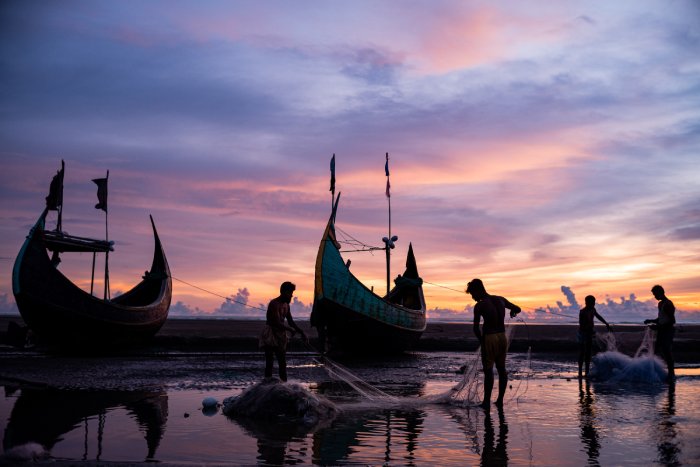 Men untangle fishing nets as primitive boats lie still on the shore during sunset.