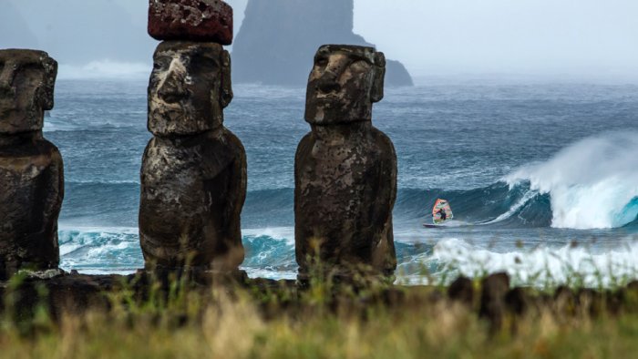 A windsurfer rides the waves on Rapa Pui with Moai statues in the foreground.