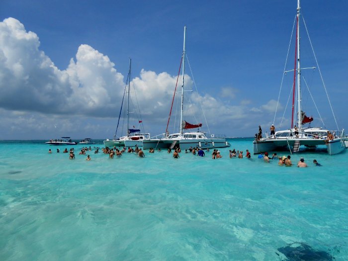 People standing in shallow tropical ocean waters next to moored catamaran sailboats.