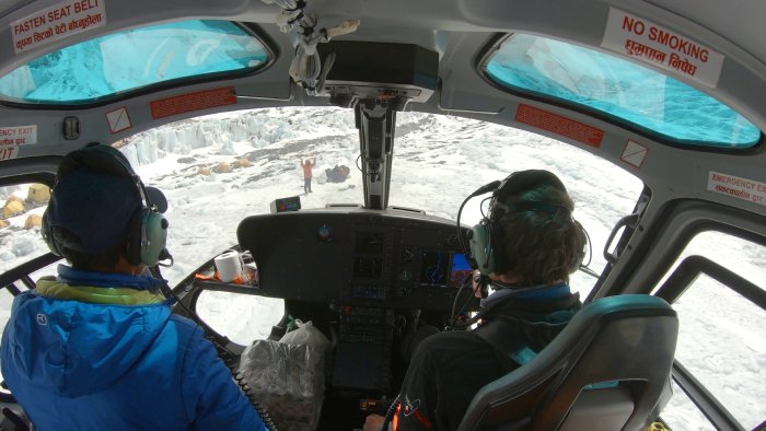 Inside the cockpit of a helicopter, looking over the shoulder of two pilots, out at a man waving the aircraft in.