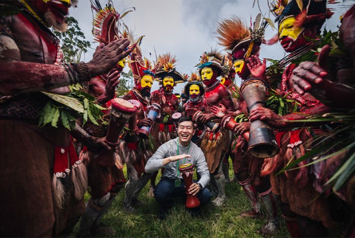 An man hits a drum as native Papua New Guinean men dressed in traditional dress dance around him.