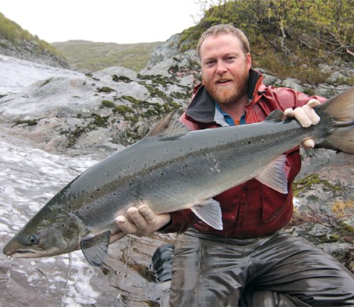 A man holds up a salmon after catching it in a rushing river.