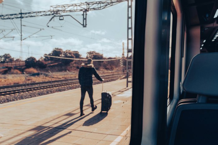 A man walks outside of a train, pulling his suitcase. The sun is low and casts long shadows on the walkway.