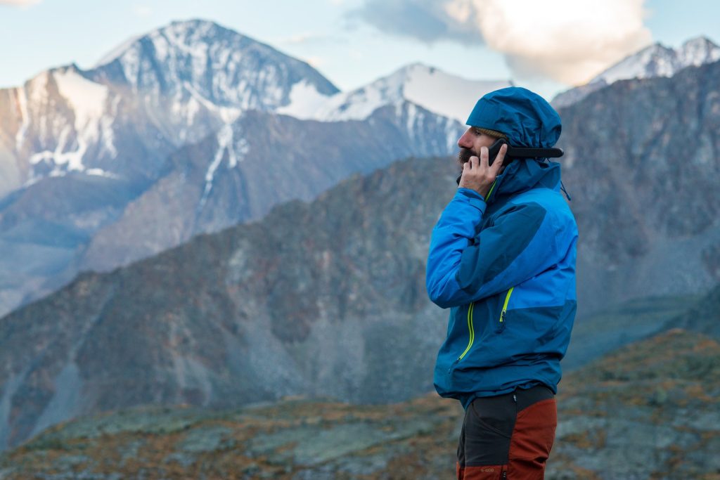 A man wearing a blue jacket and maroon technical pants uses his satellite phone in the high mountains.