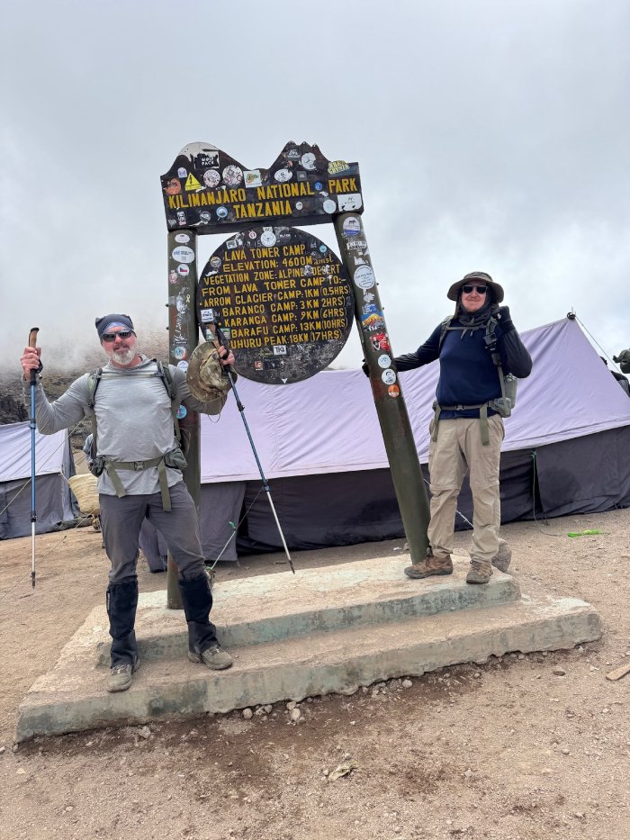 Two climbers pose at base camp beneath the Kilimanjaro sign.