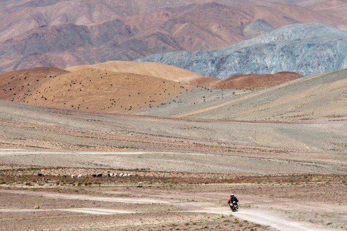 A motorcycle drives through a barren, rocky, desert landscape.