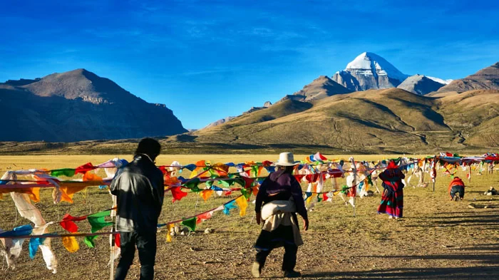 Tibetanos caminan con banderas de oración por las llanuras bajo el monte Kailash, bajo un cielo azul despejado.