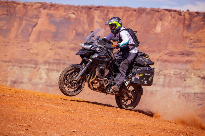 A touring motorcyclist lifts his bike's front-wheel off the desert ground, off-road in a rocky, red canyon.
