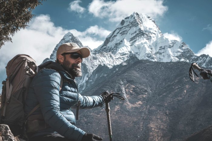 A man with sunglasses, beard, hat, winter jacket, backpack, and trekking poles sits and admires the view of the mountains behind him.