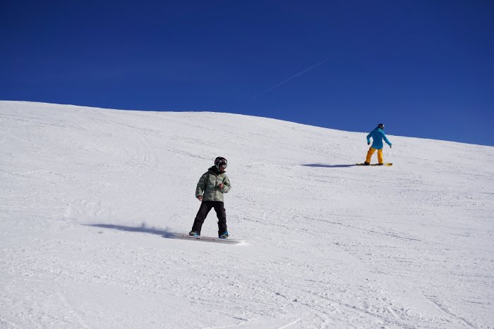 Dos snowboarders bajan por una pista nevada bajo un cielo azul.