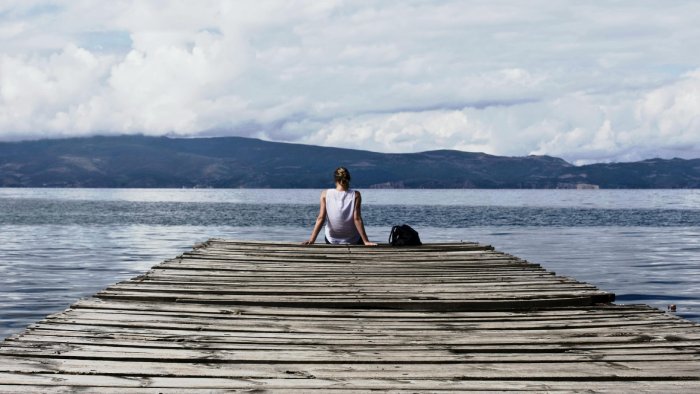 A woman sits at the end of a dock with her bag, looking out across the bay to rolling hills on the other side.