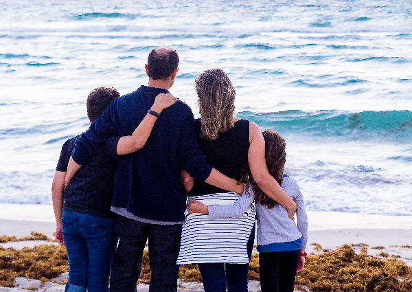 A family of four embrace each other and stand next to the water as they look out at the ocean.