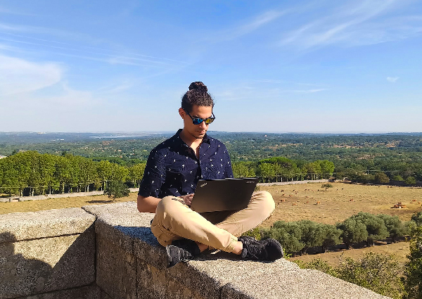 A young adult wearing sunglasses sits cross-legged on top of an old wall while working on his laptop outside.