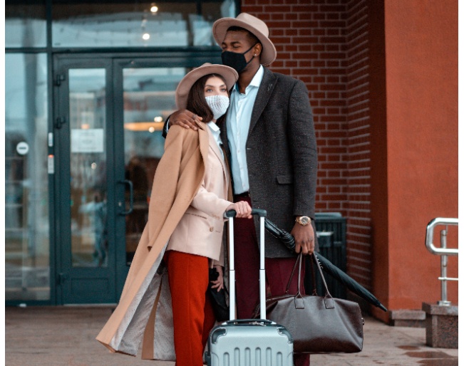 A well-dressed couple wearing masks and carrying luggage outside the doors of their hotel building.