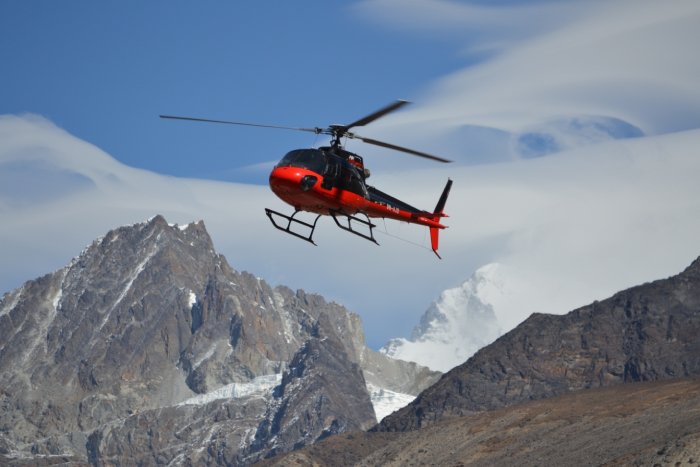 A red and black helicopter approaches a landing on a high-altitude mountain under partly cloudy skies.