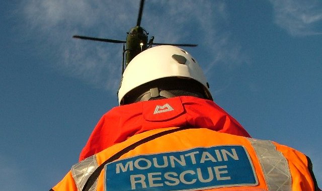 Un miembro del equipo de rescate de montaña con chaqueta naranja y casco blanco espera en el suelo bajo un helicóptero que se aproxima.