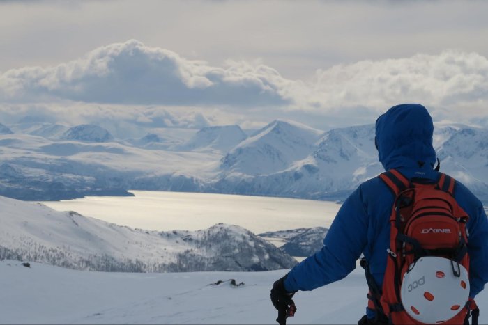 A skier in a blue jacket and backpack with helmet hanging pauses to observe the view while on a ski tour.
