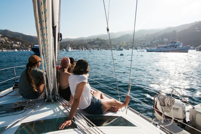 Four women sit on the deck of a sailboat in harbor under the sunlight.