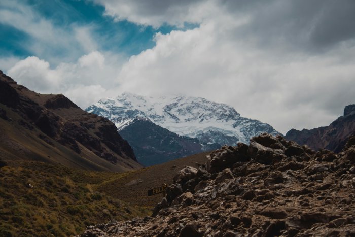 A snowy mountain towers over smaller brown hills under a cloudy sky.