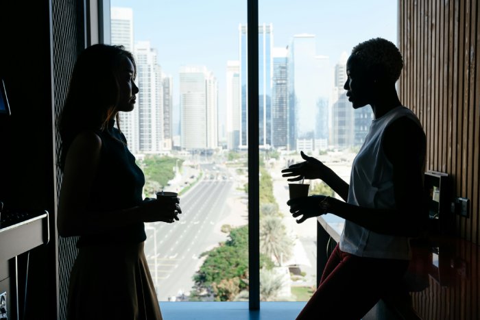 Two multicultural women talk in an office building in front of a window.