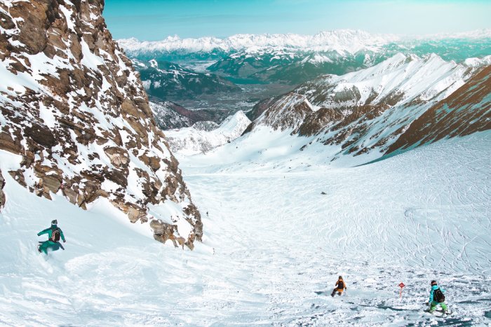Three snowboarders ride down an open snow field with cliffs on the side.