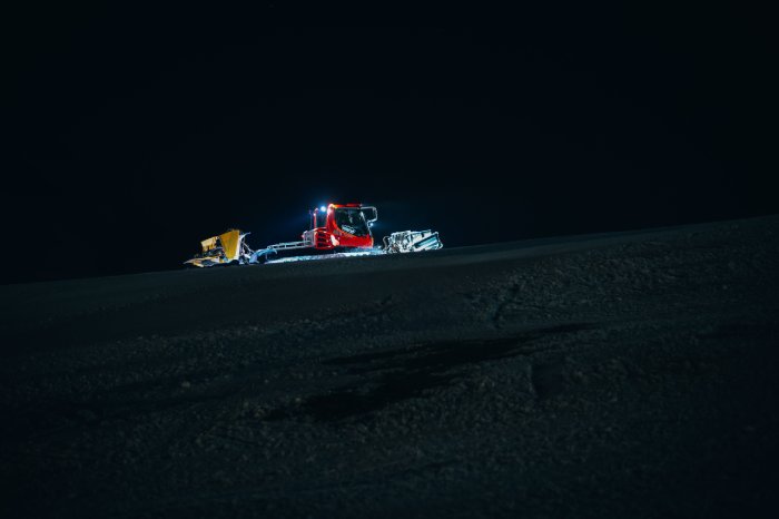 A snowcat climbs a ski slope at night to groom the snow.