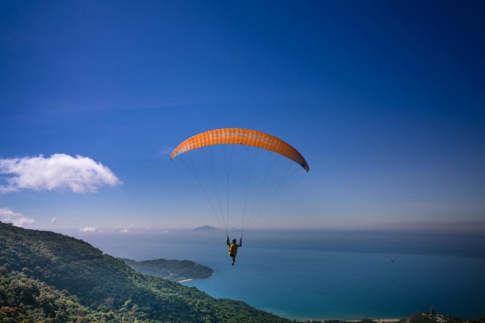 A paraglider high above the green land and blue ocean under a bright sky.