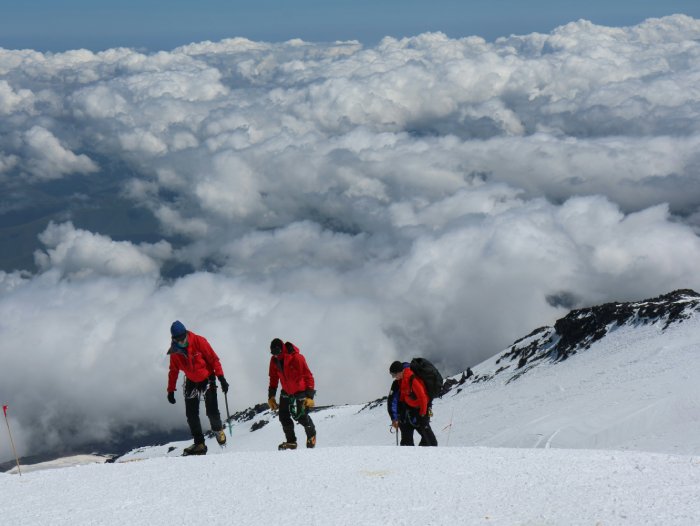 Three climbers in red jackets climb high above the clouds in the snow on a mountain.