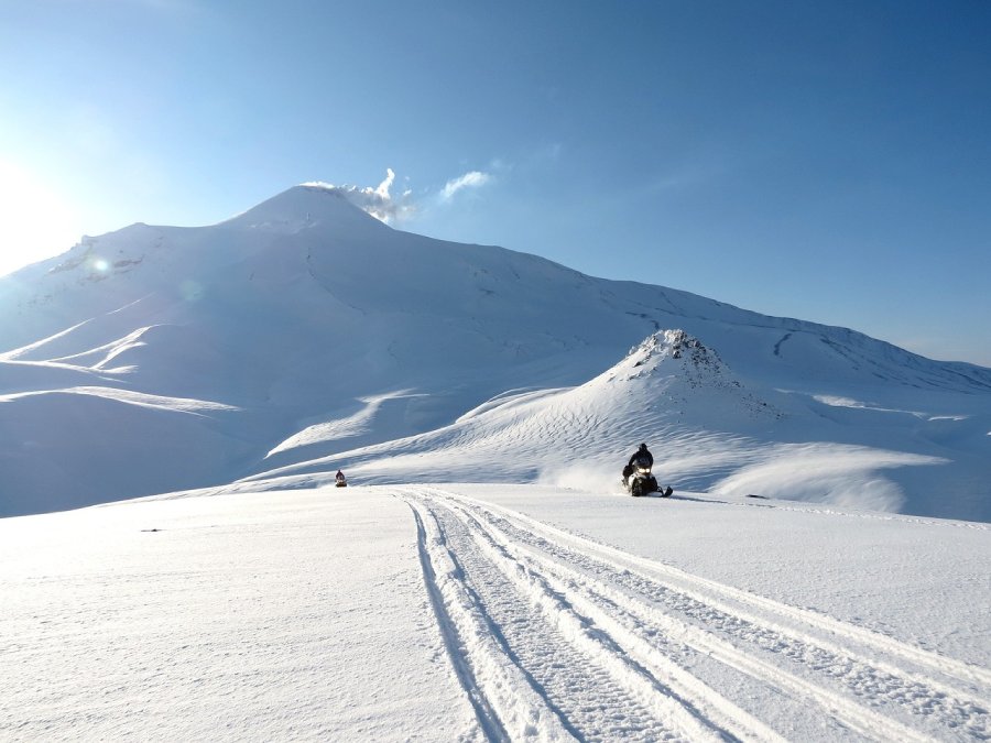 Two snowmobilers ride in deep snow under blue skies.