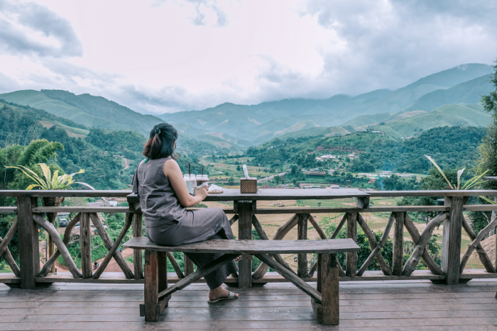A woman sits on a bench on a deck eating, looking out over a rice paddy in Thailand.