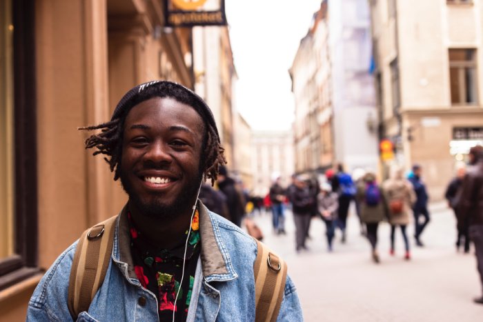 A smiling young man walks along a crowded city street with a backpack and headphones on.